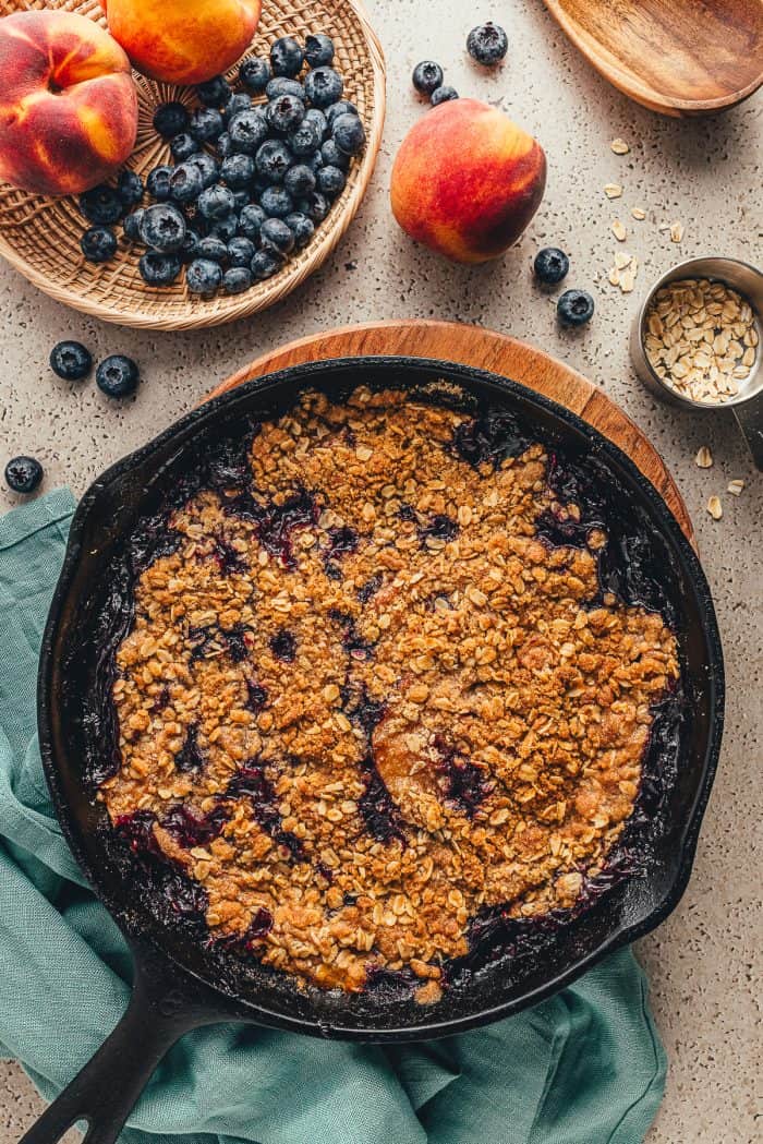 An image of a skillet with a crumble in it and a bowl of blueberries  and peaches and a teal cloth. 