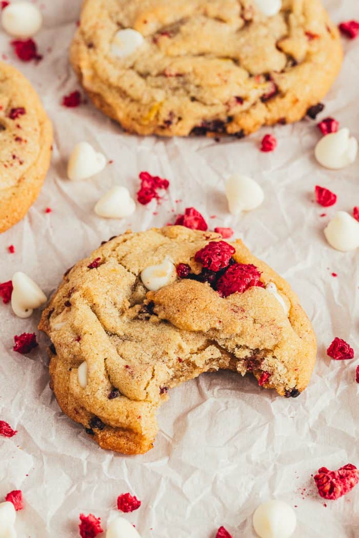 A raspberry white chocolate cookie with a bite taken out of it and white chocolate chips and freeze-dried raspberries scattered around.