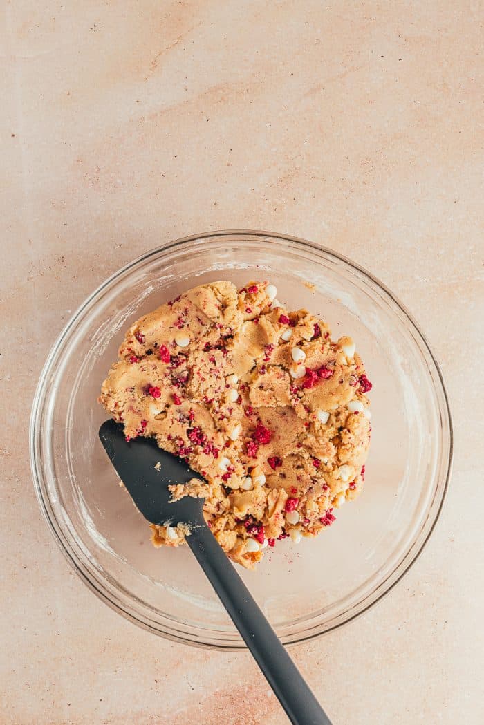 A glass bowl with cookie batter and freeze-dried raspberries and a spatula.