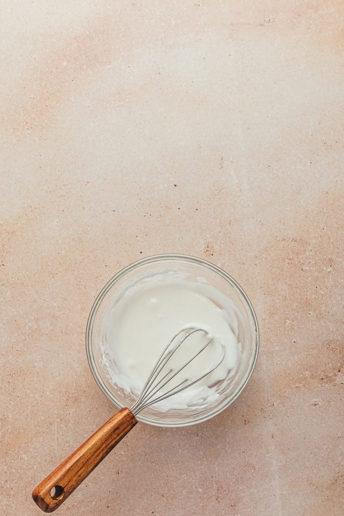 A glass bowl with icing in it and a whisk.