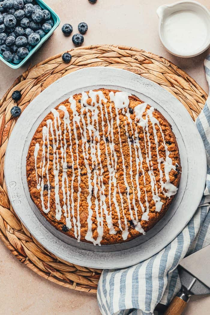 The whole blueberry sour cream coffee cake with icing drizzled all over it on a large white plate.