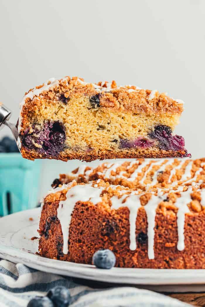 A slice of blueberry coffee cake on a cake server with the whole cake in the background.