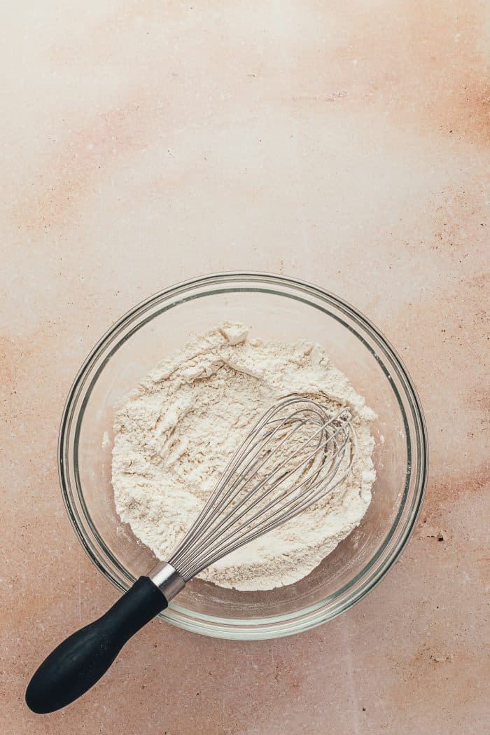 A glass bowl with flour in it and a whisk.