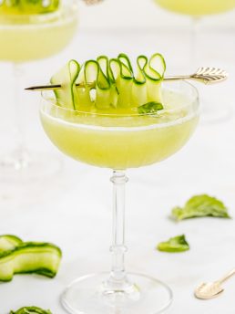 A gimlet garnished with a cucumber ribbon with basil in the background.