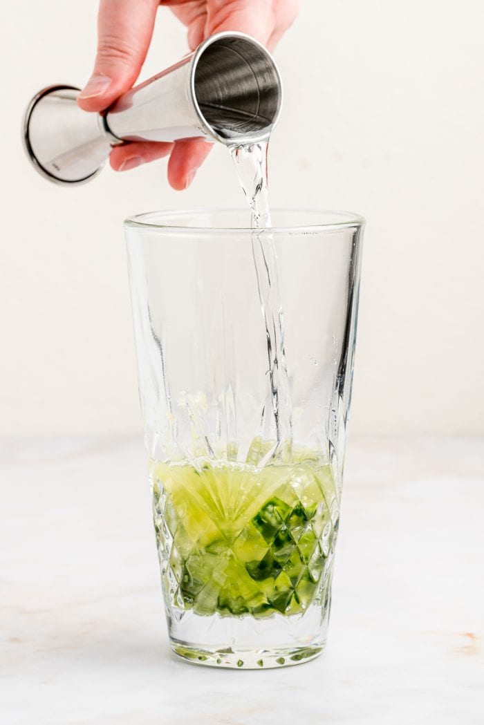A glass with muddled cucumber and gin being poured into it.