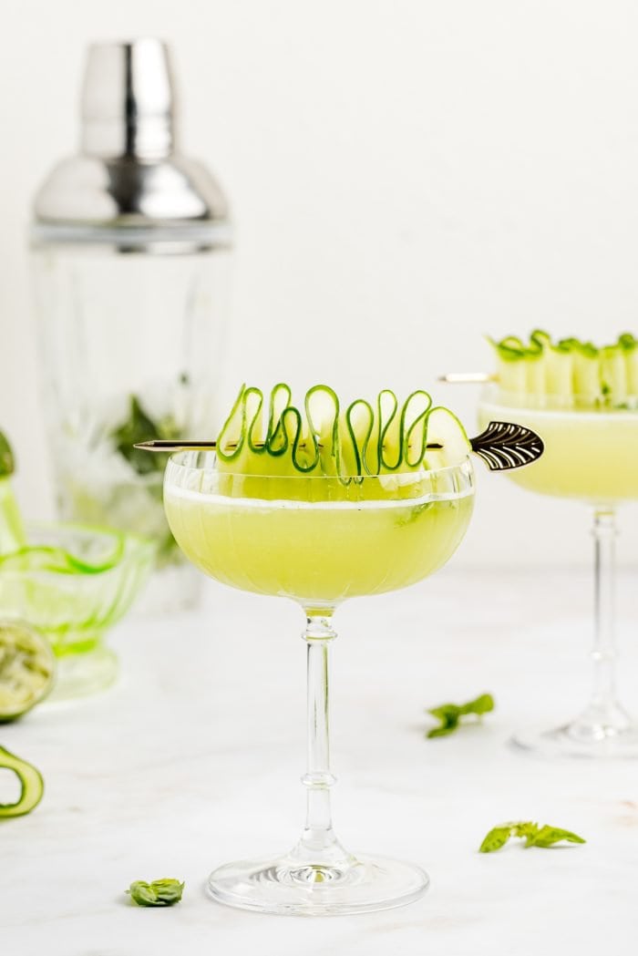 A gimlet garnished with a cucumber ribbon with basil in the background.