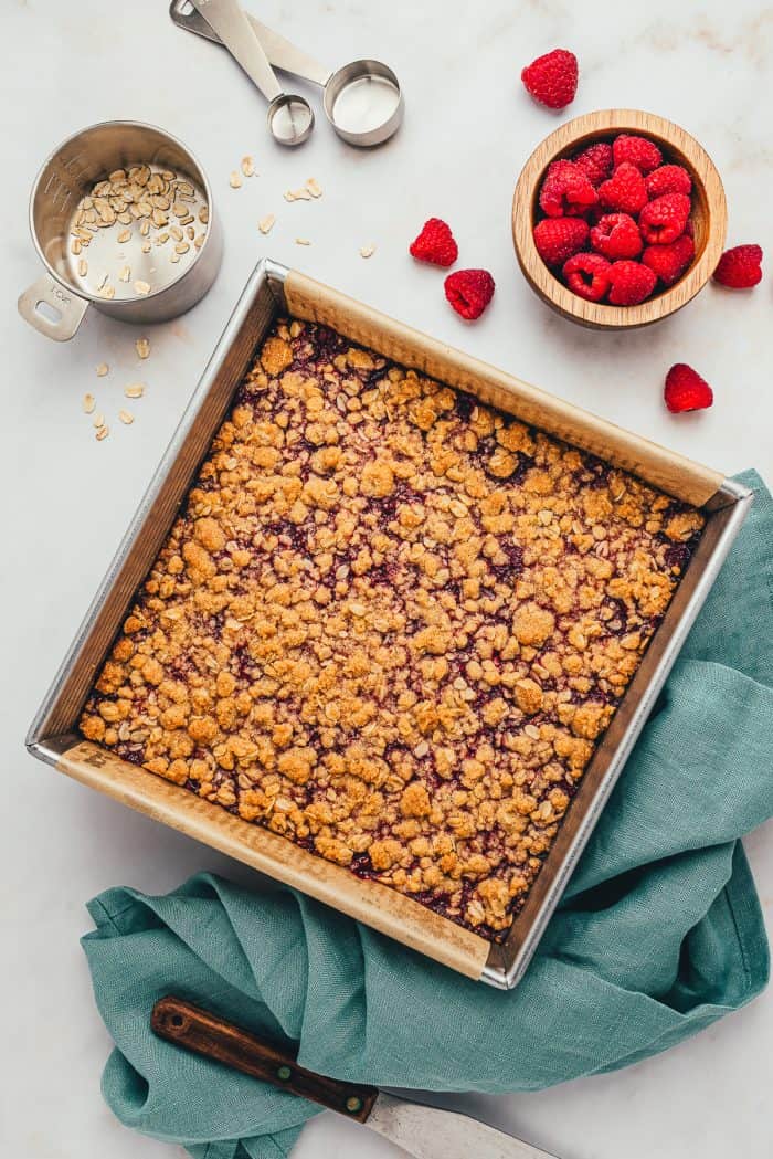 The baked raspberry crumble bars with a teal colored towel and fresh raspberries scattered.