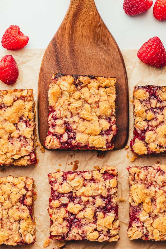 Six raspberry bars with one being removed on a wooden spatula with fresh raspberries scattered around.