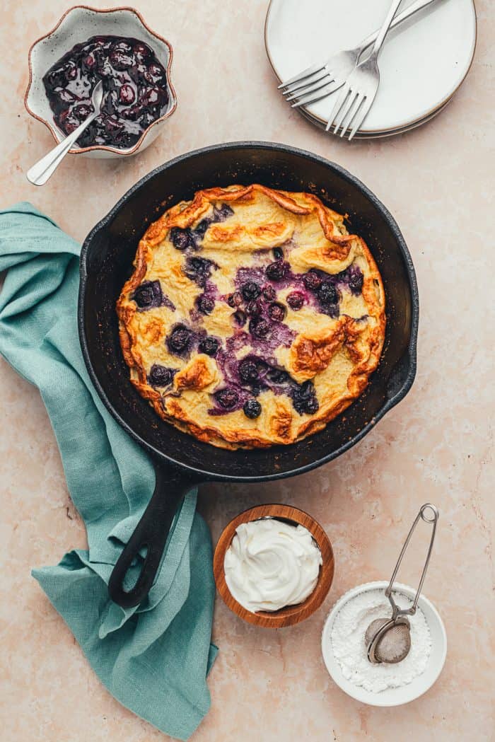 A skillet with a baked blueberry Dutch baby pancake. Bowls of garnishes and blueberry compote.
