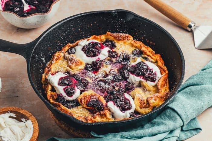 A skillet with a baked blueberry lemon Dutch baby pancake that is garnished with whipped cream and blueberry compote.
