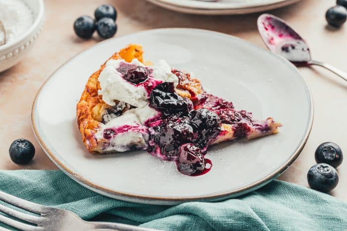 A plate with a slice of blueberry lemon Dutch baby pancake with a dollop of whipped cream and blueberry compote.