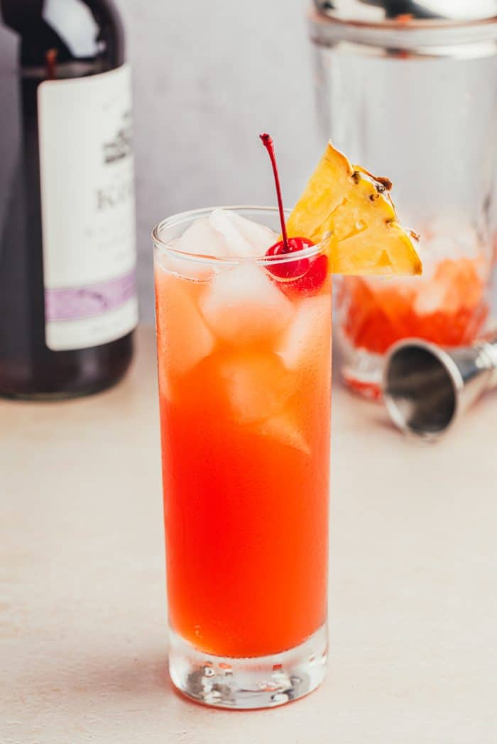 A glass filled with Planter's Punch and decorated with a maraschino cherry and a slice of pineapple with a bottle of rum and a cocktail shaker in the background.