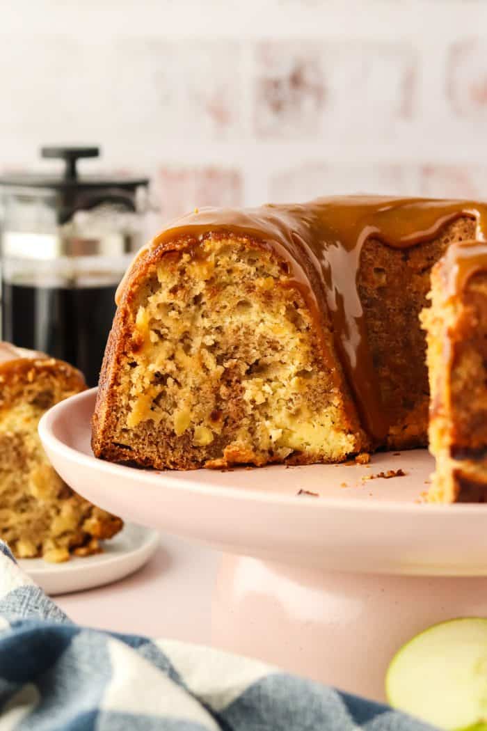 A caramel apple Bundt cake with slices removed from it on a white cake plate.