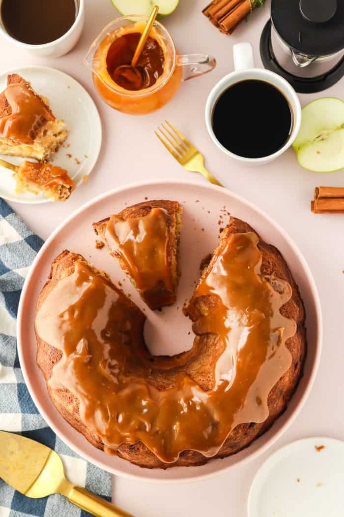 An apple Bundt cake with caramel sauce with slices cut out of it on a cake stand. 