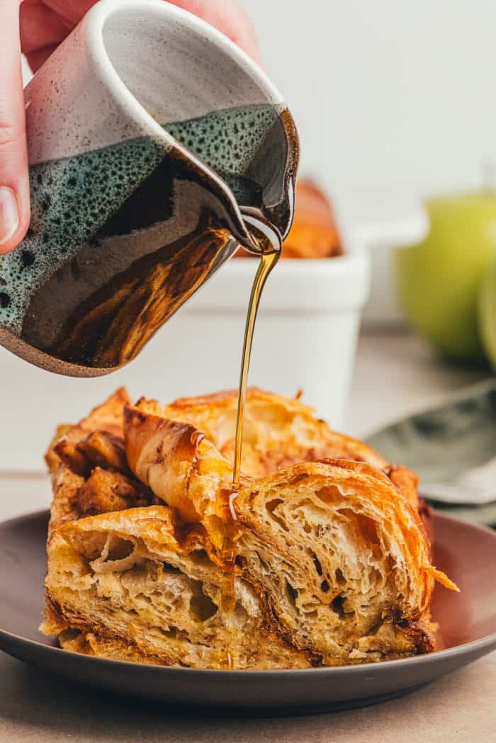 A slice of apple french toast bake on a brown colored plate with a bottle of maple syrup being poured over it.