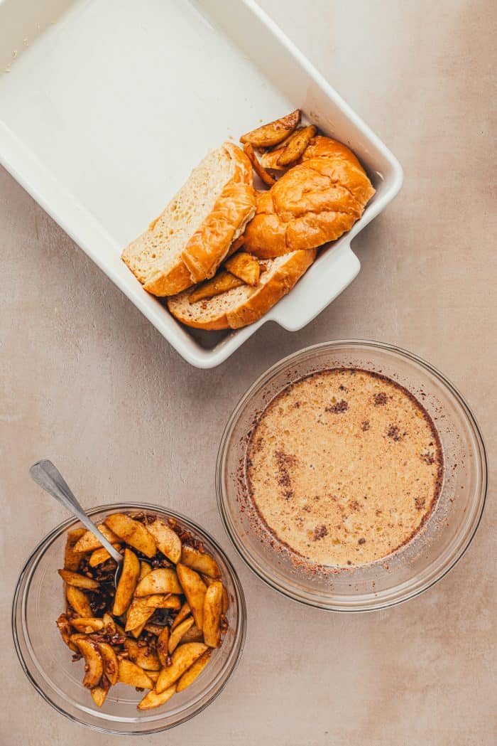 A white baking dish with freshly dipped croissants and a bowl of apple pie filling and french toast egg mixture.