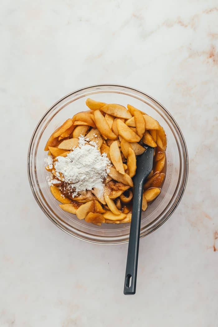 The apple pie filling in a glass bowl with cornstarch sprinkled over it and a black spatula. 