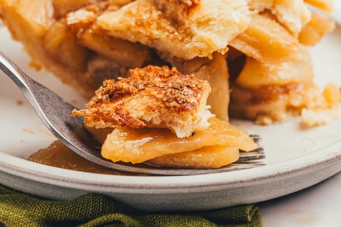 A fork with a bite of apple pie on it on a white plate with the apple pie slice behind it.