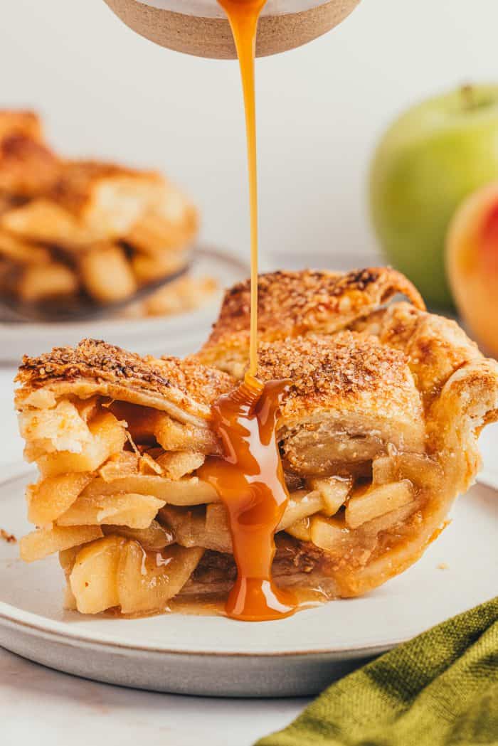 Caramel sauce being poured over a slice of apple pie on a white plate.