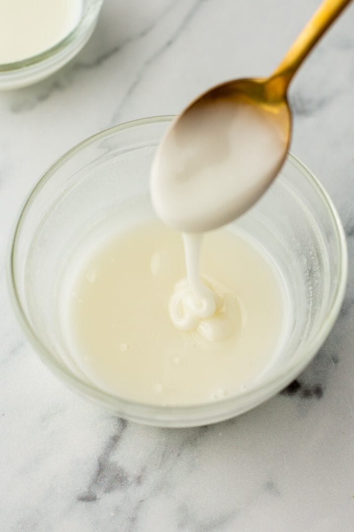 A glass bowl of icing with a spoon drizzling the icing into the bowl.