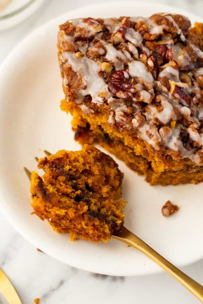 A white plate with a slice of pumpkin coffee cake with a fork slicing into it.