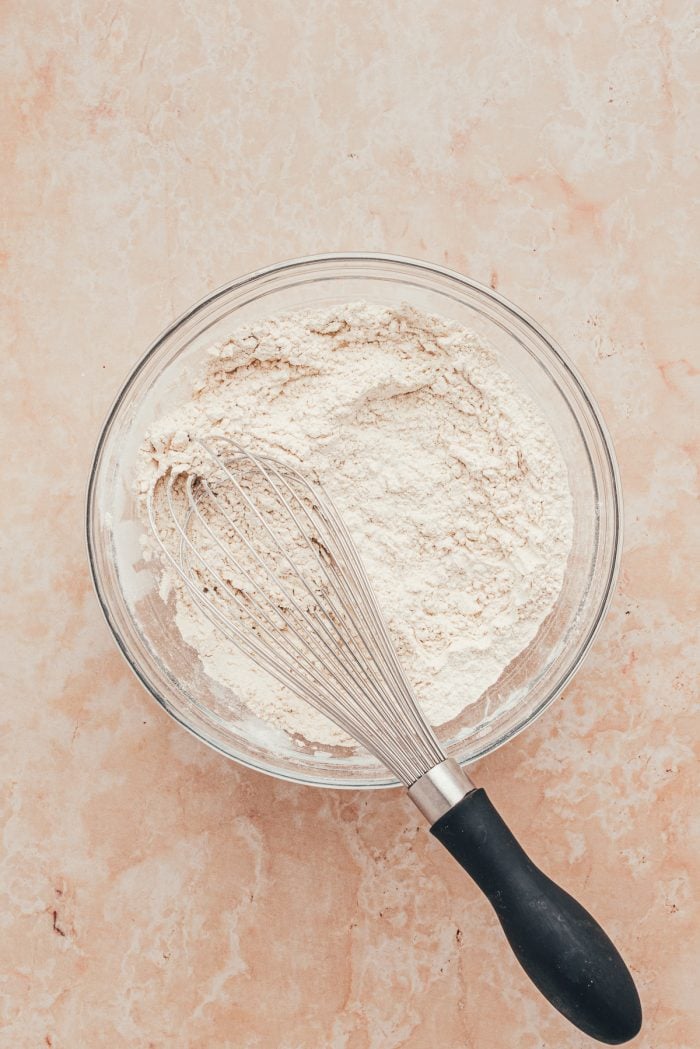 A glass bowl with flour and spices whisked together and a whisk.