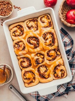 A white baking dish with baked apple pecan cinnamon rolls with caramel sauce.