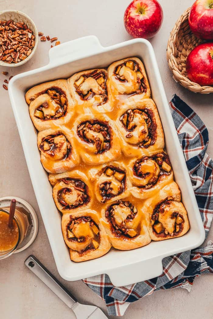 A white baking dish with baked apple pecan cinnamon rolls with caramel sauce.