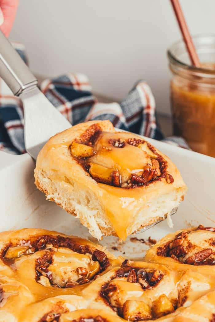 A spatula lifting a cinnamon roll out of a white baking dish.