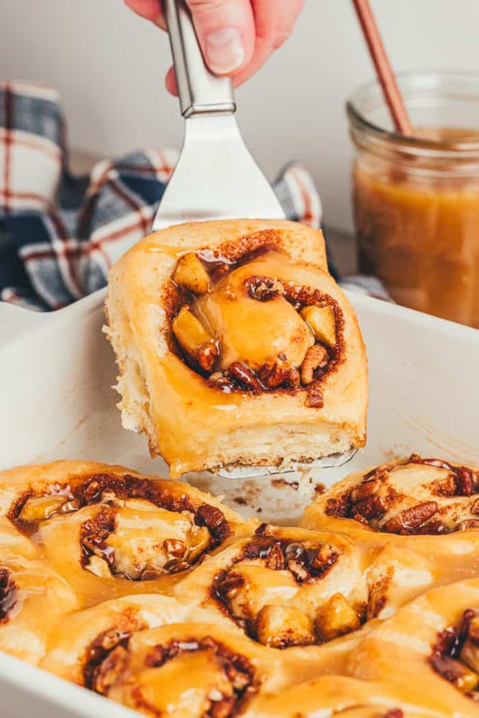 A spatula lifting a cinnamon roll out of a white baking dish.