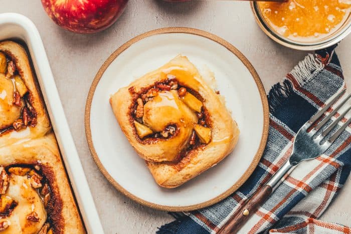 A white plate with a cinnamon roll and apples in the background.