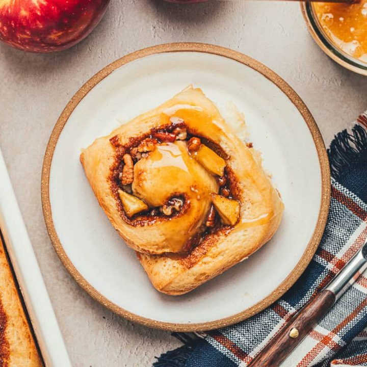 A white plate with a cinnamon roll and apples in the background.