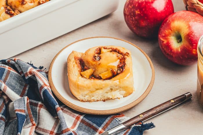 A white plate with a cinnamon roll and apples in the background.