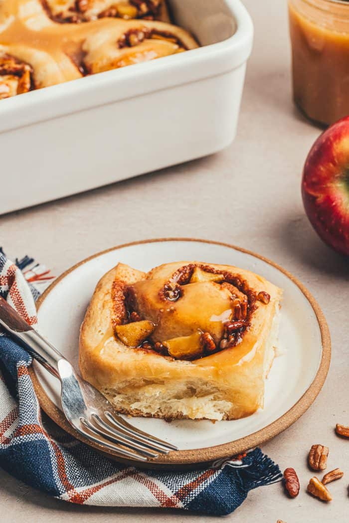 A white plate with a cinnamon roll and apples in the background.