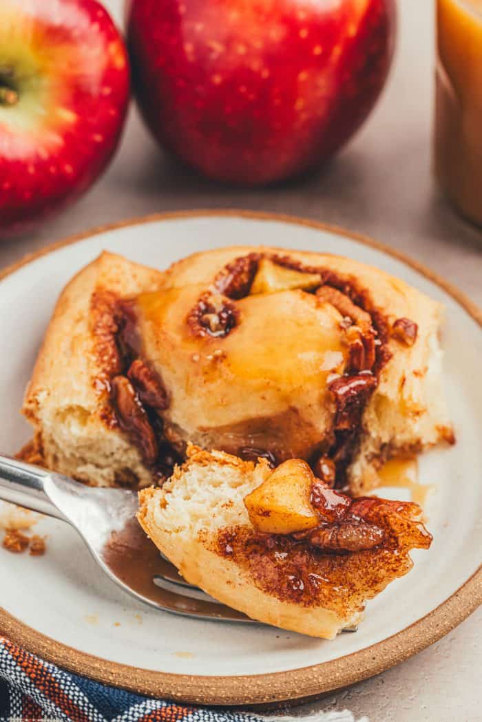 A closeup of a caramel apple cinnamon roll that is pulled apart and has a fork scooping up a piece on a white plate.