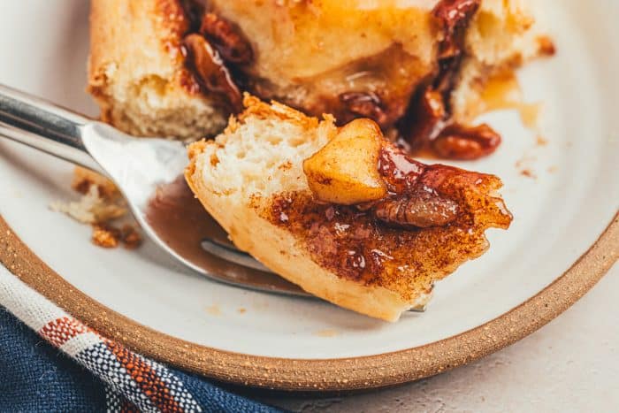 A closeup of a caramel apple cinnamon roll that is pulled apart and has a fork scooping up a piece on a white plate.