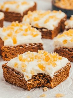 The gingerbread bars with frosting and candied ginger cut into squares and one has a bite taken out of it.