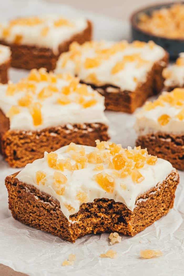 The gingerbread bars with frosting and candied ginger cut into squares and one has a bite taken out of it.