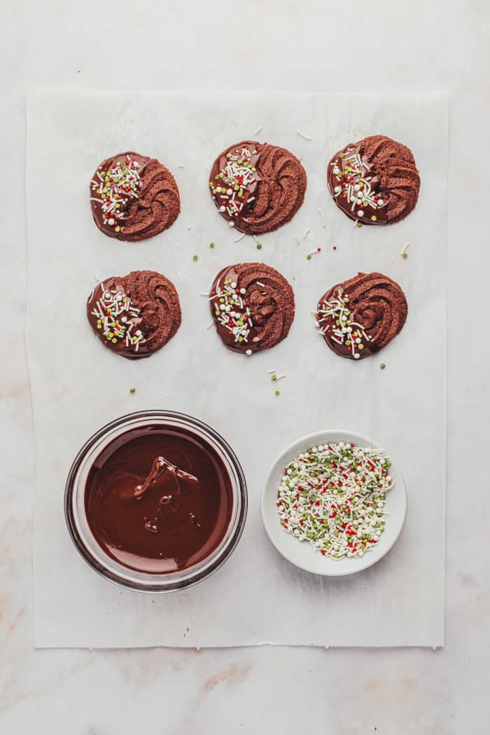 The chocolate dipped cookies on parchment paper with a bowl of melted chocolate and a bowl of sprinkles. 