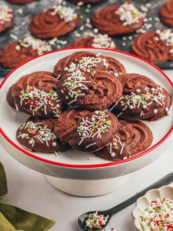 A cake platter with a pile of orange chocolate cookies and a spoon of sprinkles.