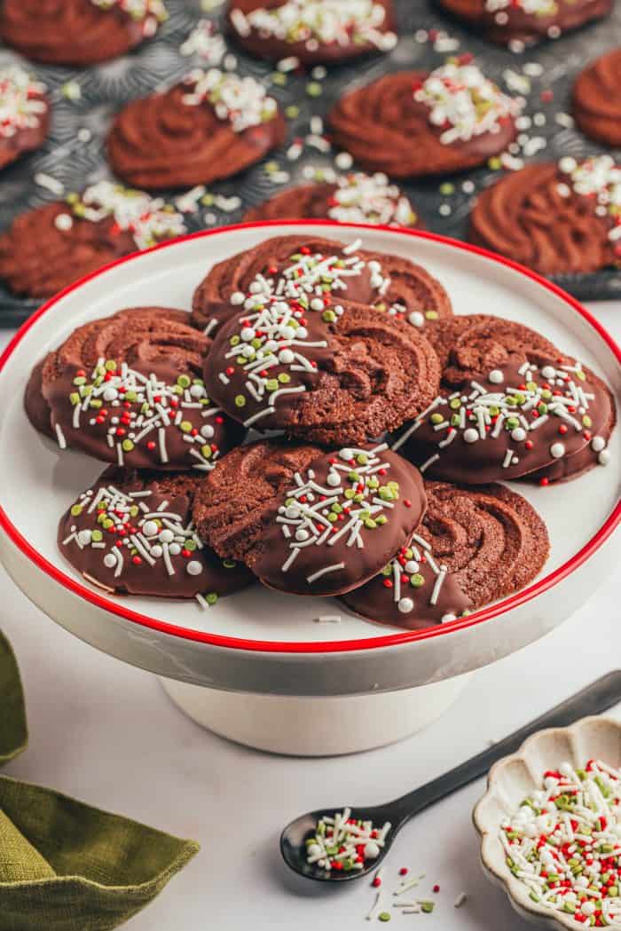 A cake platter with a pile of orange chocolate cookies and a spoon of sprinkles.