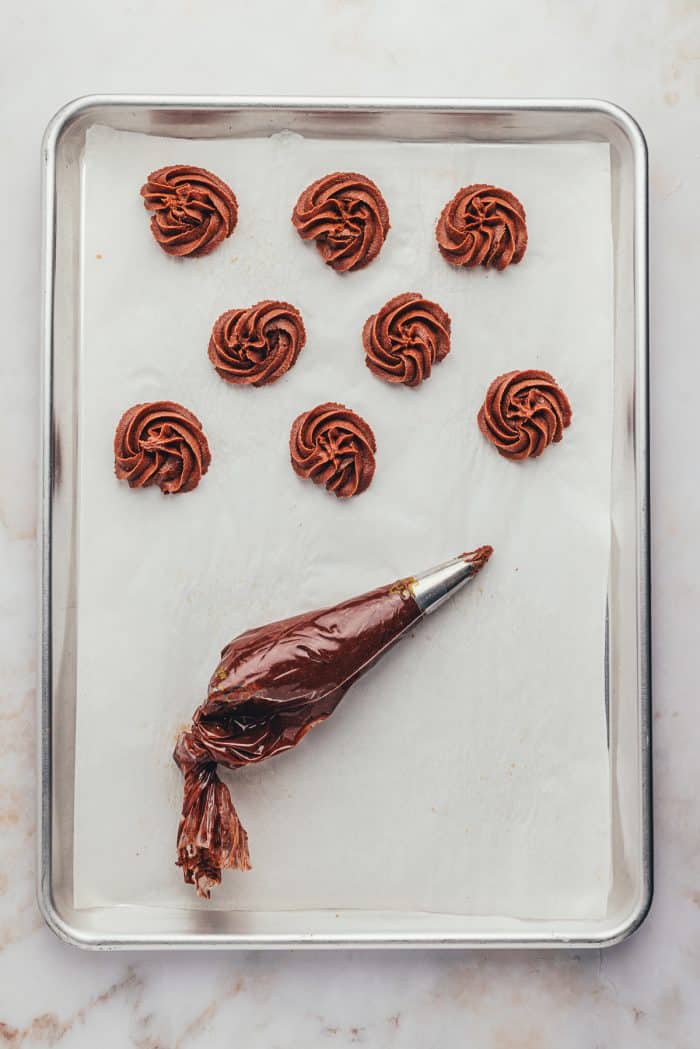 A piping bag filled with cookie dough and the cookies piped onto a parchment-lined baking tray.