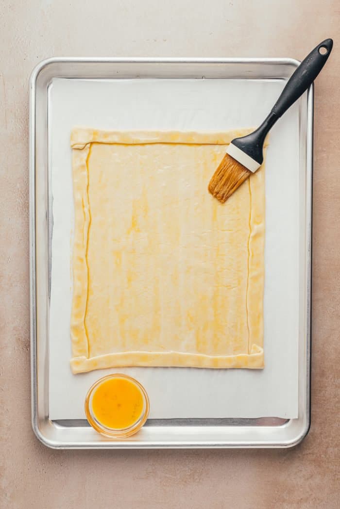 Puff pastry rolled out on a baking sheet with the edges turned in to make a crust, with egg wash and a pastry brush.