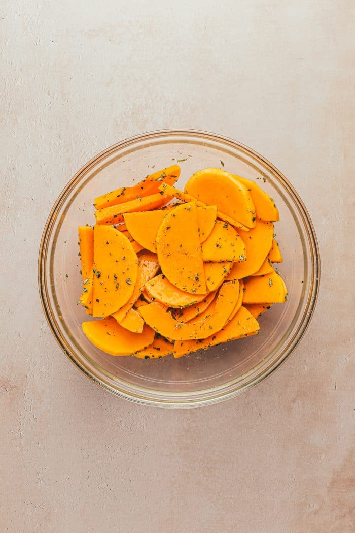 A glass bowl with sliced butternut squash mixed with herbs and olive oil.