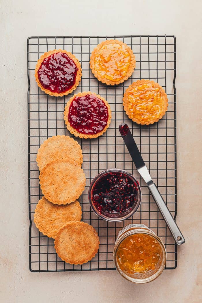 Baked Linzer cookies with jam spread on them and a knife and two bowls of jam on a cooling rack.