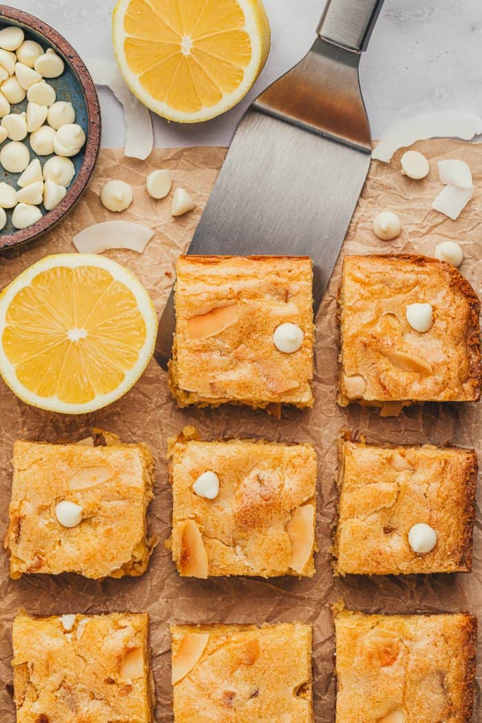 Lemon blondies with a spatula going under one blondie and half of a lemon with a bowl of white chocolate chips.