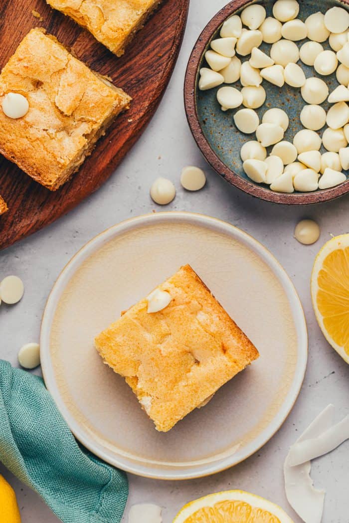 A lemon blondie on a small plate with a bowl of white chocolate chips.