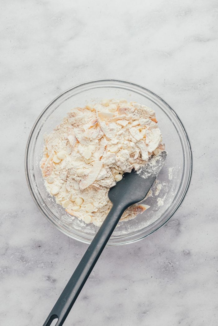 A glass bowl with flour and coconut flakes mixed together and a spatula.