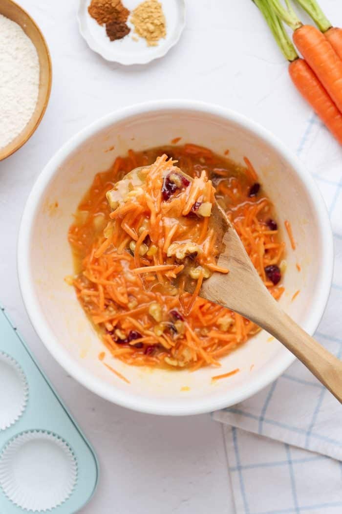 A white mixing bowl with all of the ingredients to make the carrot cake cupcakes batter and a spoon with some of the batter on it.