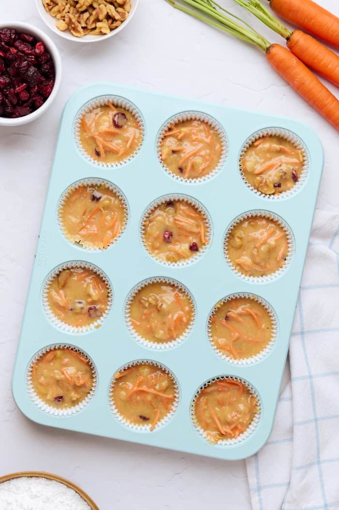 A blue cupcake tray with cupcake batter in it and carrots in the background.